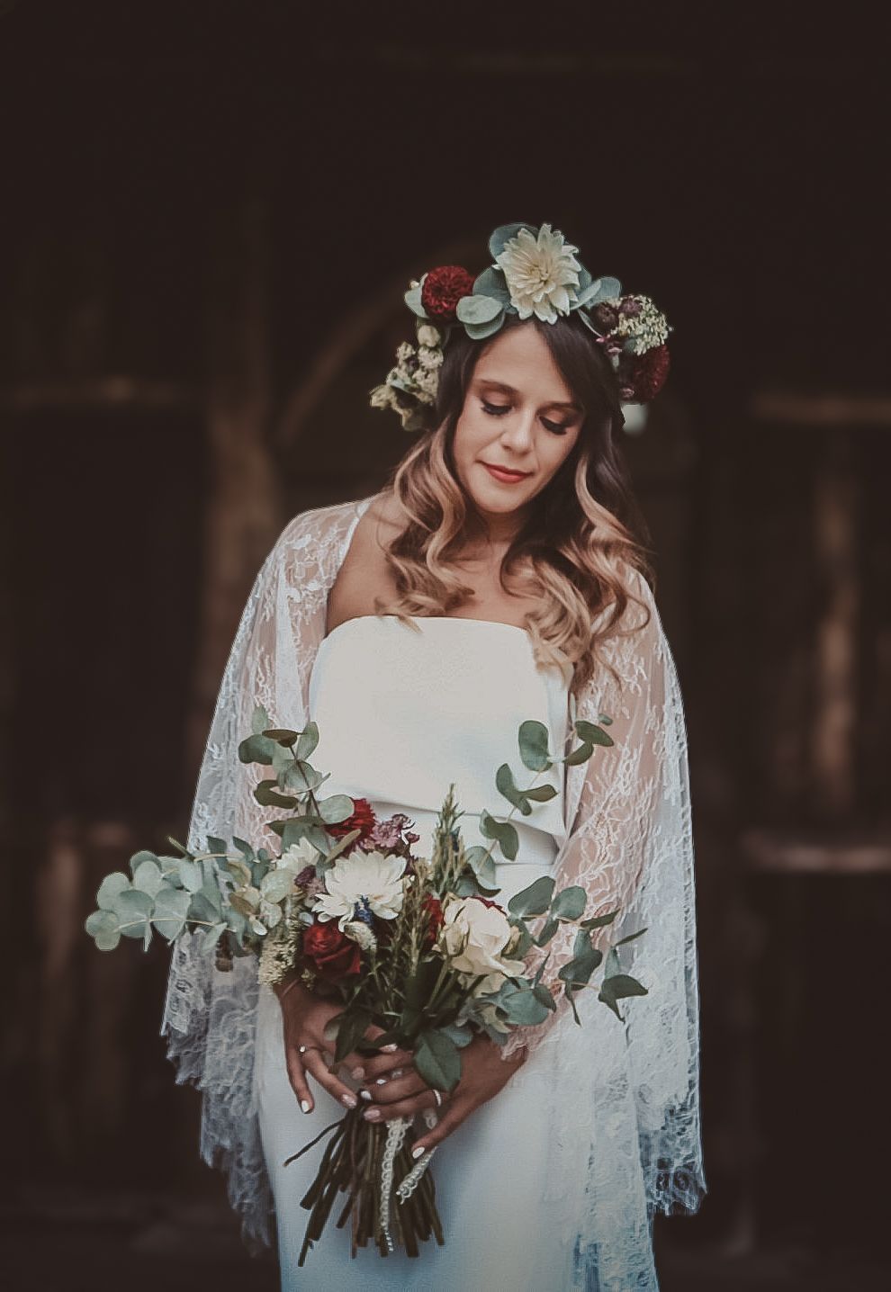 A bride in a white lace dress and floral crown holds a romantic burgundy and eucalyptus bouquet in moody lighting.