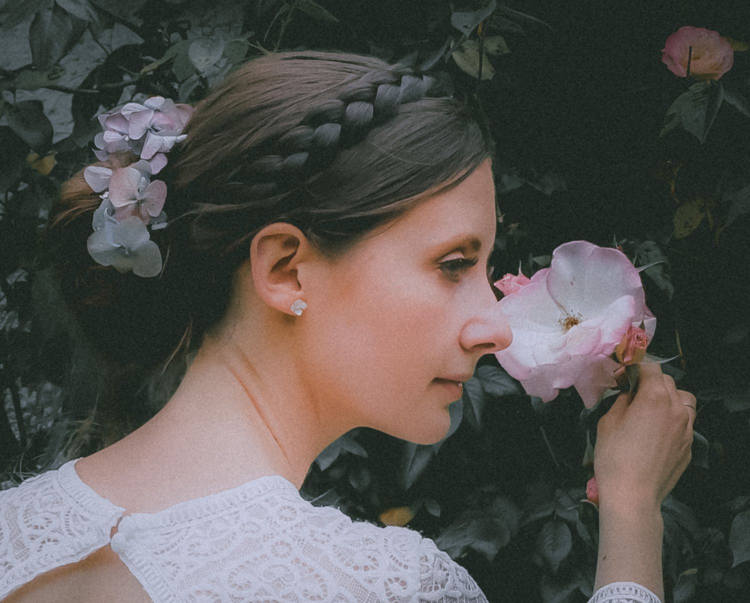 A feminine figure in white lace smells a delicate pink rose against a dark garden background.