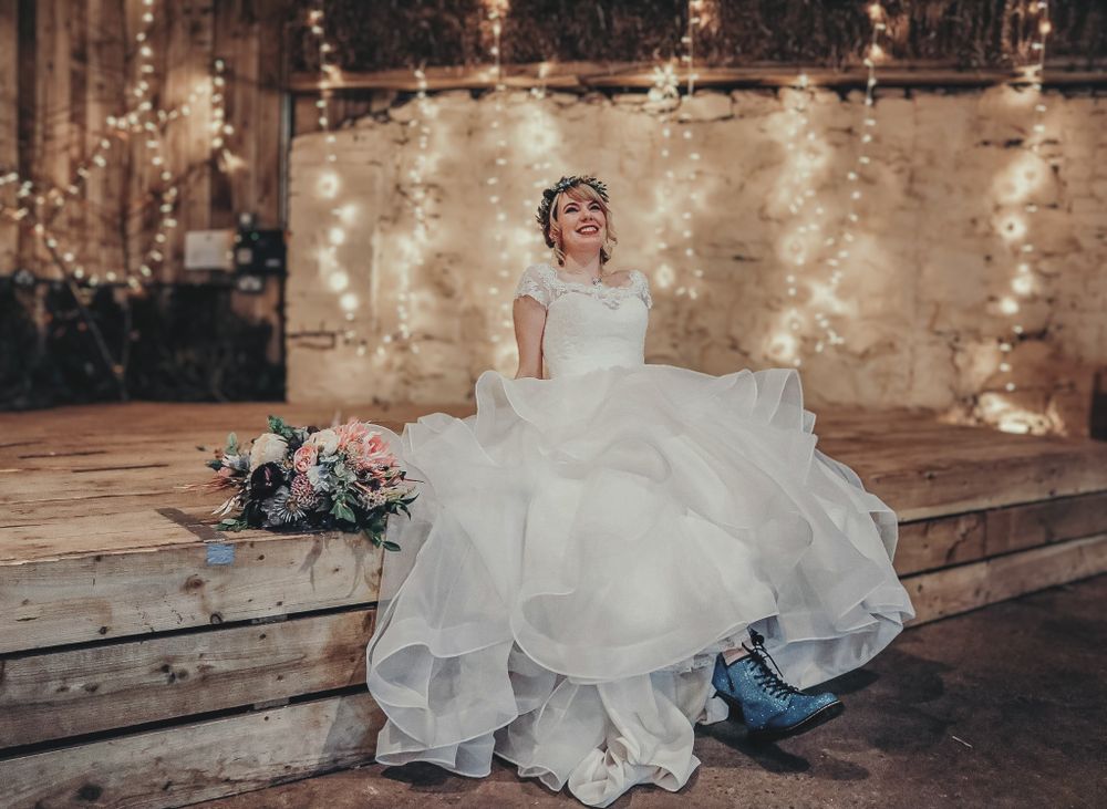 A person in a flowing white wedding dress sits on wooden steps with a bouquet and string lights in the background.