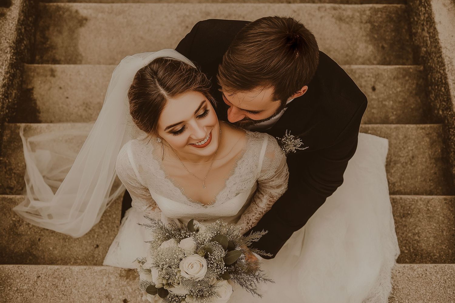 Overhead view of a bride and groom sitting on stone steps with a floral bouquet in a warm sepia-toned photograph.