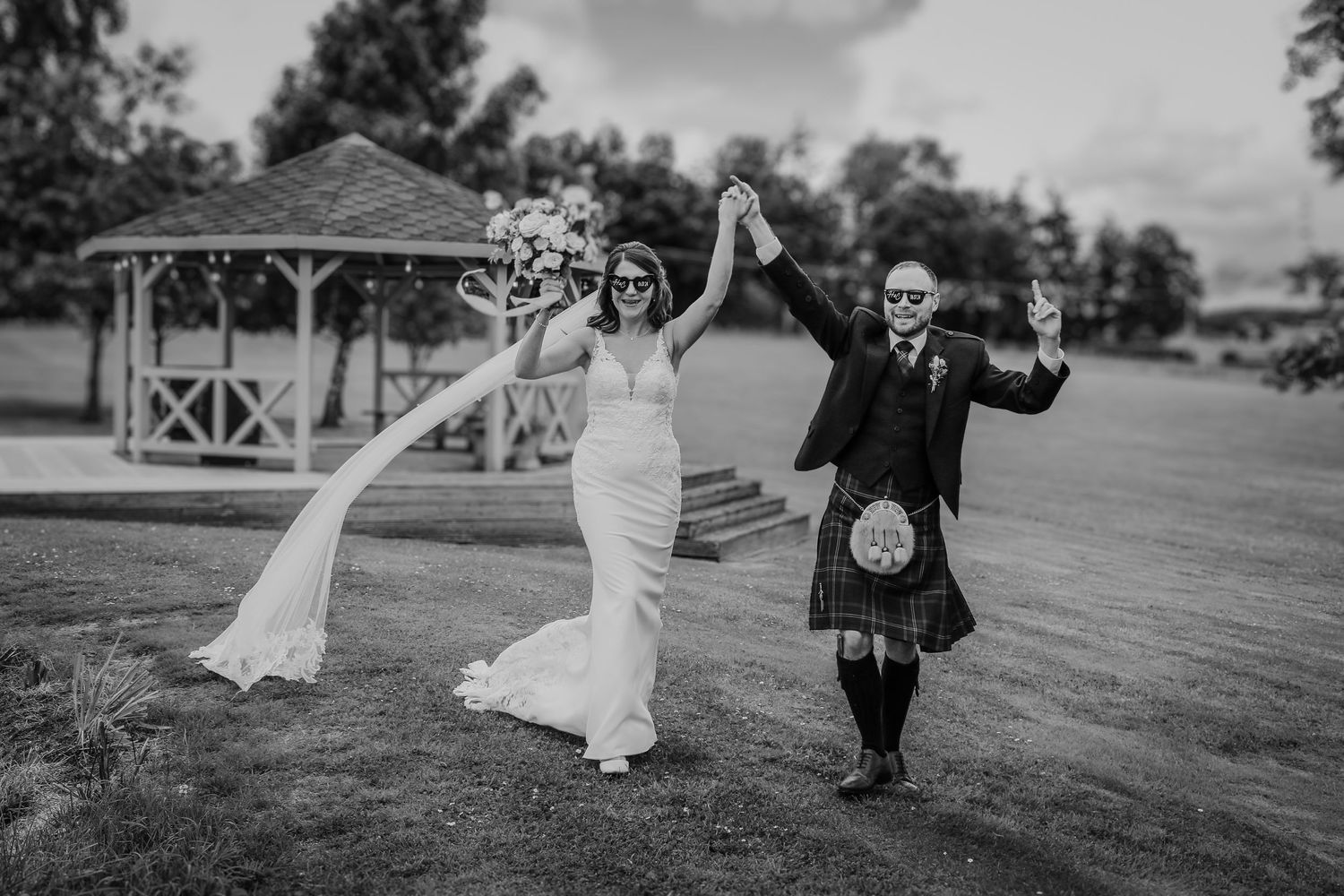 Black and white wedding photo showing couple celebrating on a lawn with a gazebo in the background.