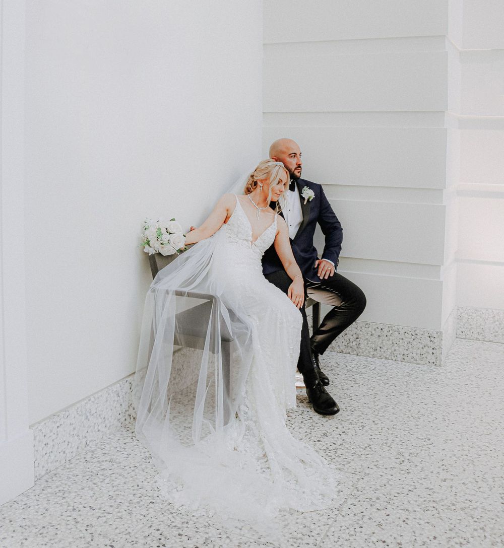 A romantic black and white wedding photo of a couple in formal attire sitting on a bench against a light wall.