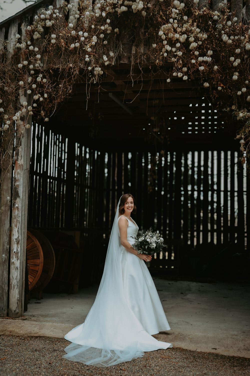 White wedding gown with flowing train photographed in a rustic barn setting with hanging florals and wooden beams.