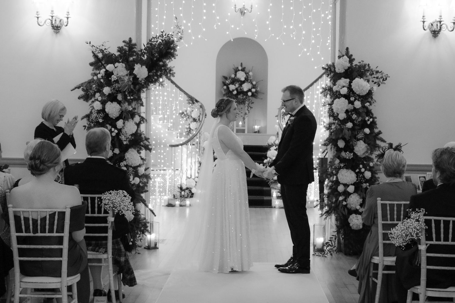 A black and white photograph of a wedding ceremony in a chapel decorated with elegant floral arrangements and wall sconces.