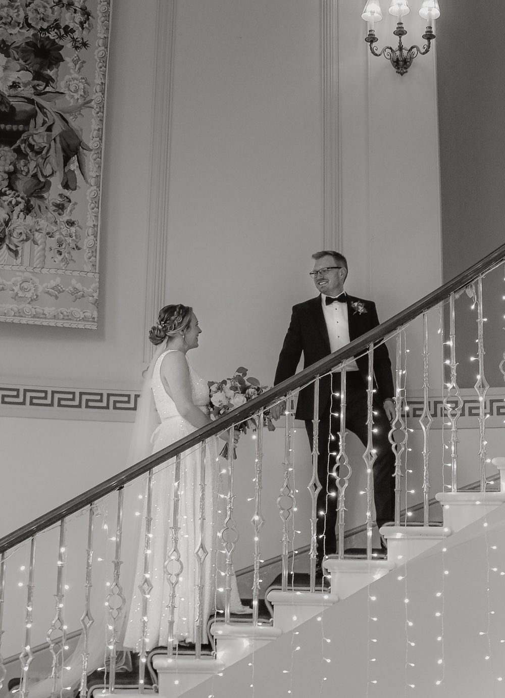 Elegant black and white wedding photo on staircase decorated with twinkling string lights and ornate Greek key molding.