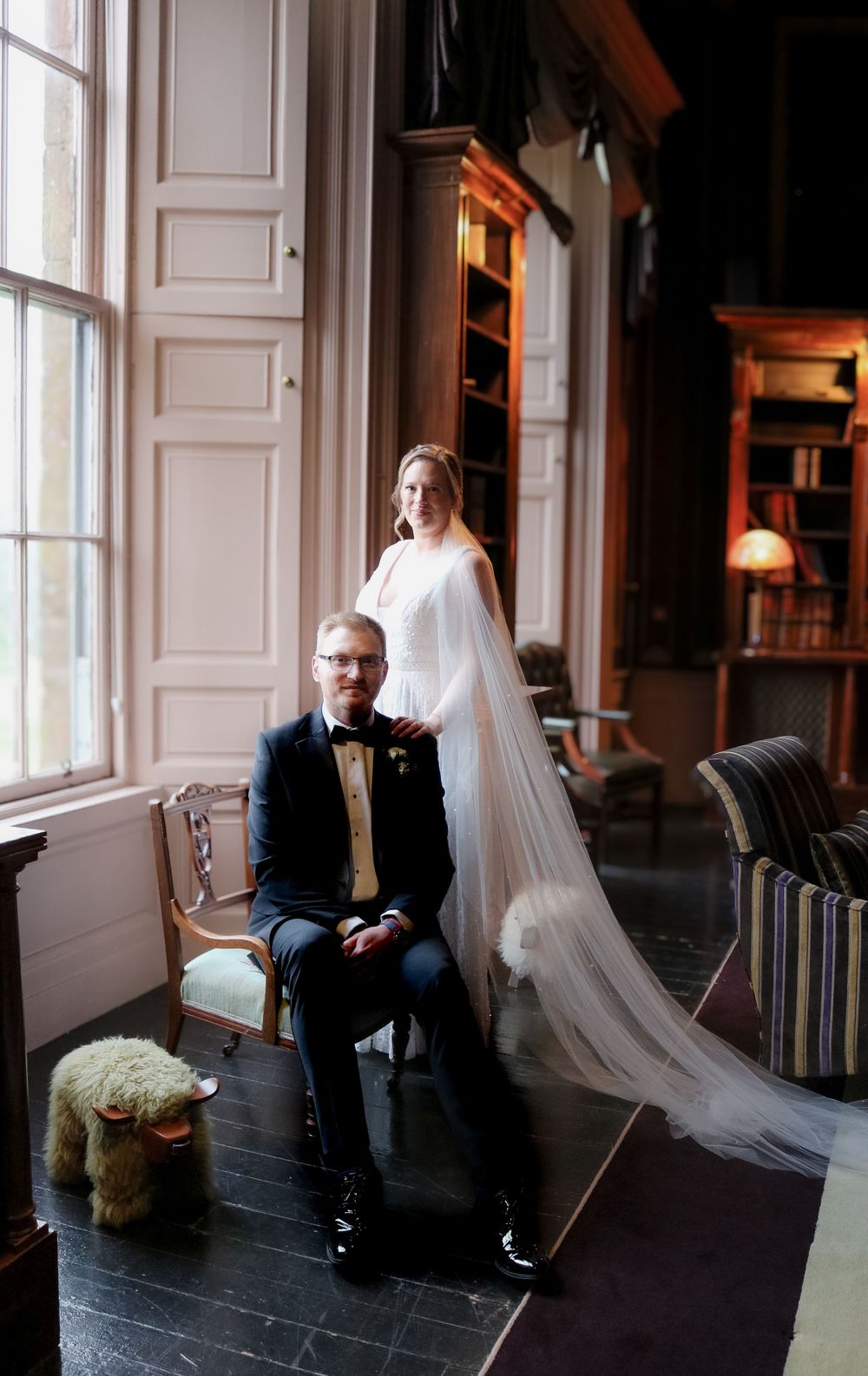 Wedding couple poses elegantly in library room with tall windows and vintage furniture.