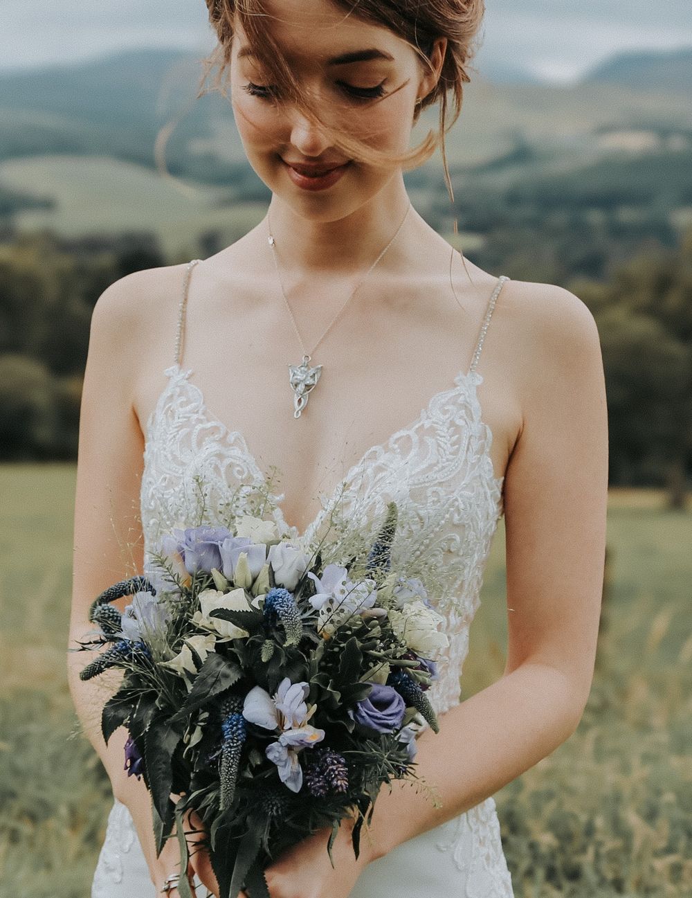 A bride in a delicate lace gown holds a beautiful bouquet of blue and white flowers in a scenic mountain setting.