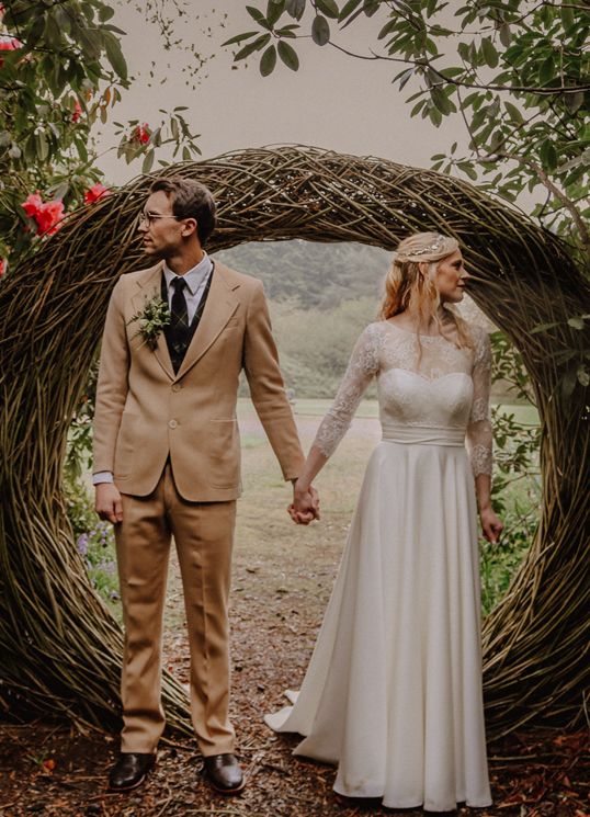 Rustic wedding ceremony arch made of woven branches with white floral gown and tan suit under natural greenery.