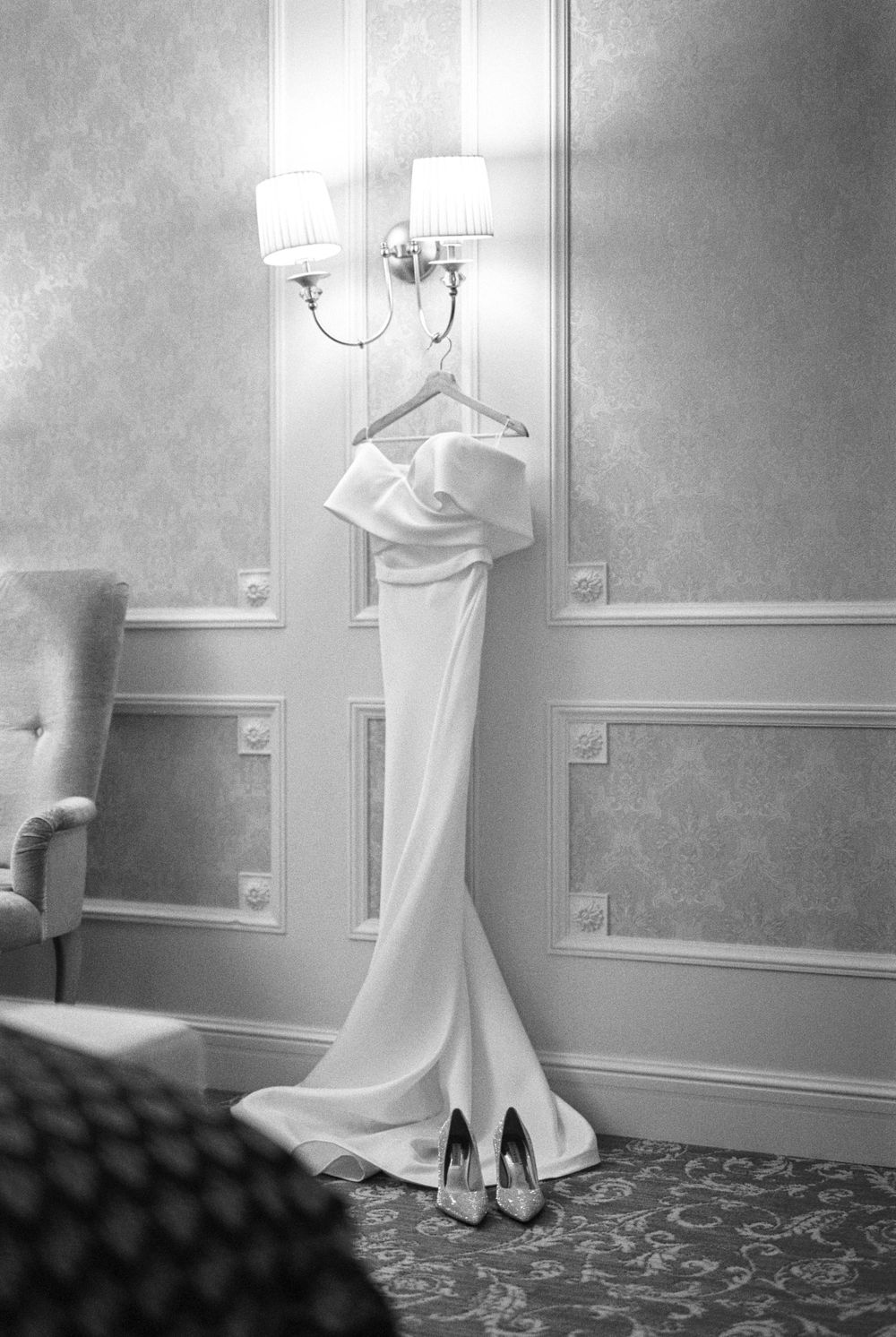 Elegant white wedding dress and silver heels displayed on hanger against wall with sconce lighting and ornate molding.